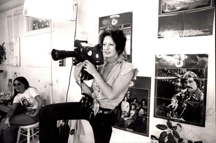 Black and white photo of a woman holding a movie camera in front of a wall of posters