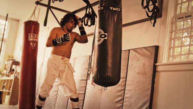 Photo of a man in white pants and no shirt with fists raised in front of a boxing bag