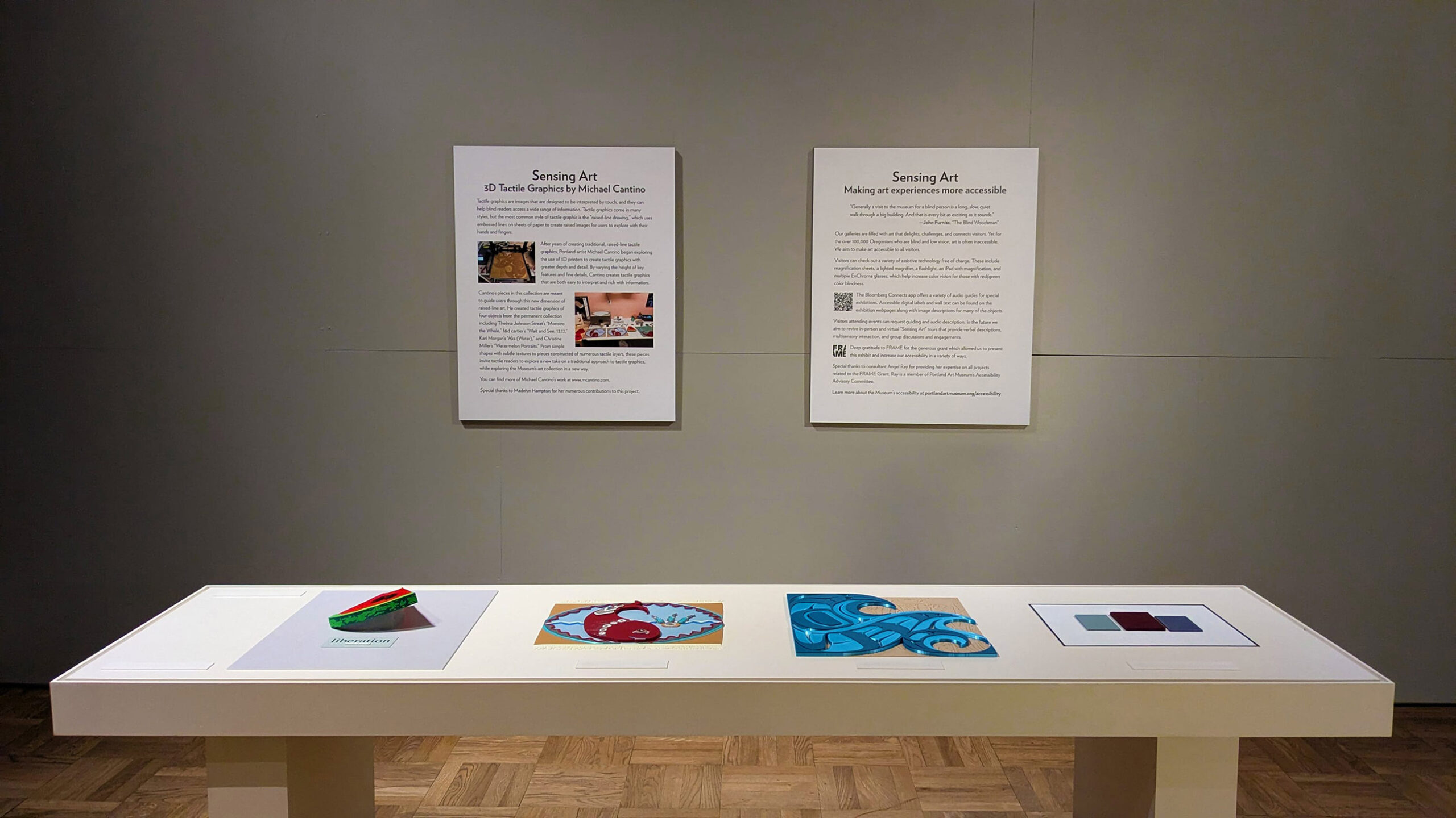 Four tactile artworks on a table with two text panels on the wall behind.