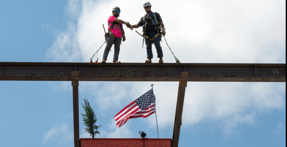 Two construction workers on top of a steel beam shaking hands