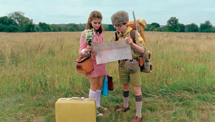Film still of two kids standing in a field of grass looking at a map. The girl is wearing a 1960s pink dress and the boy is wearing a boy scout uniform.