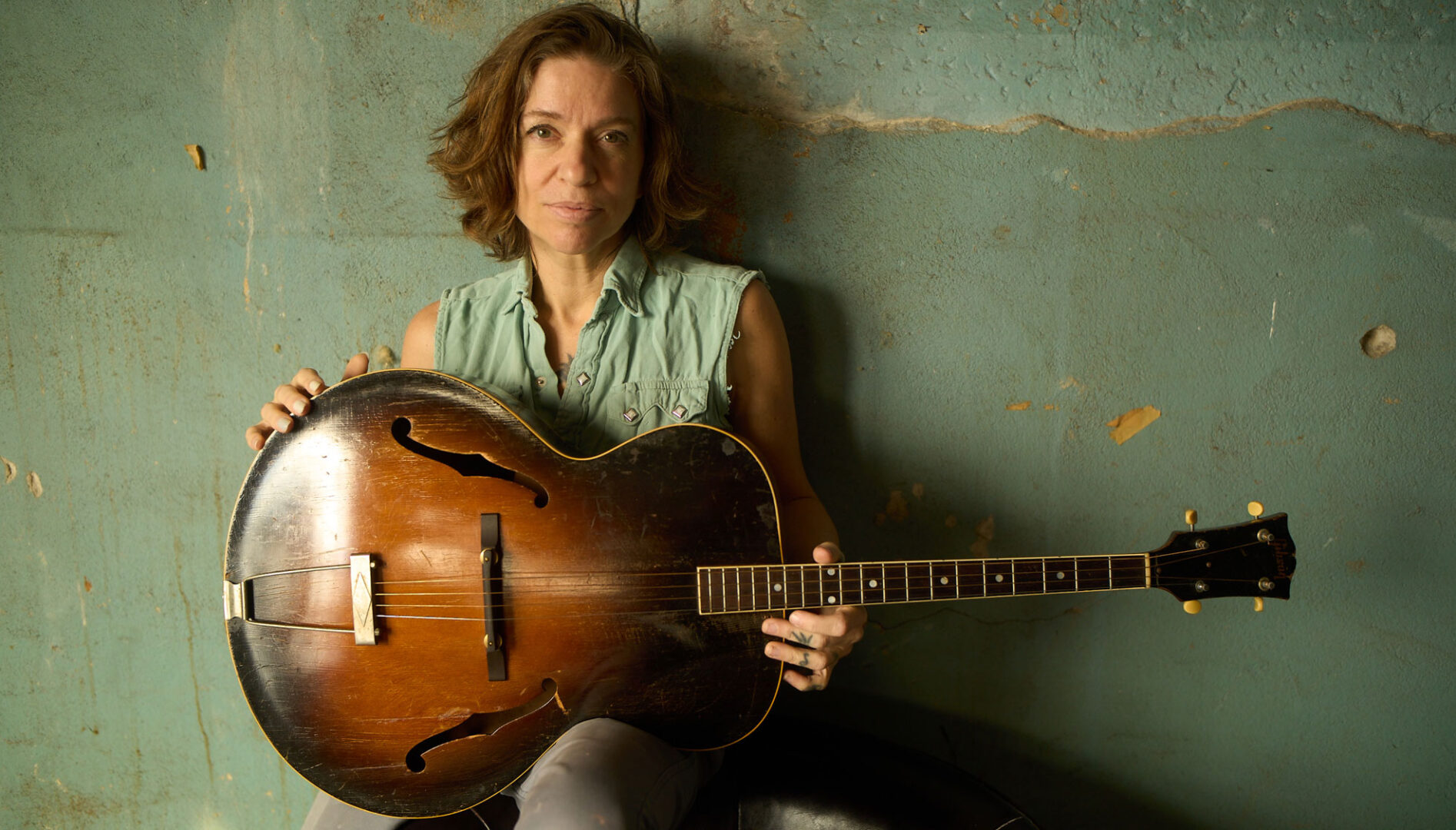 Portrait of the artist Ani DiFranco holding a guitar and sitting against a green wall