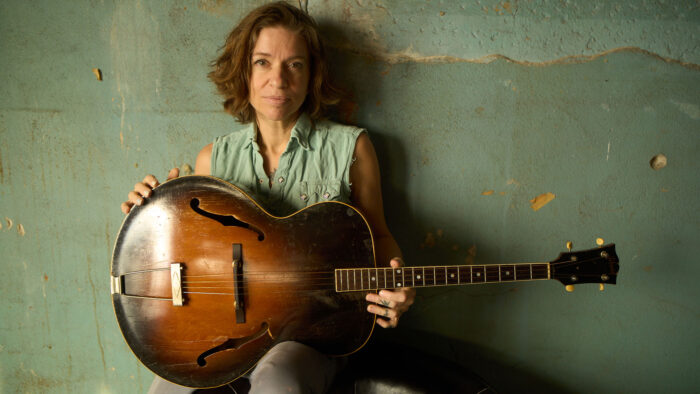 Portrait of the artist Ani DiFranco holding a guitar and sitting against a green wall