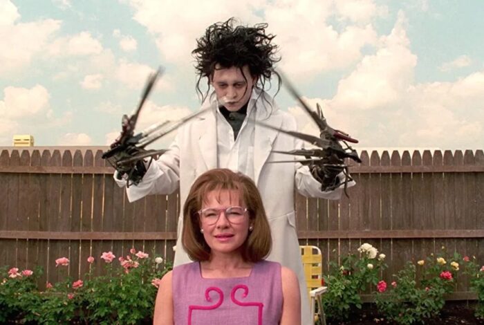 Film still of a pale man with wild black hair and a white coat who had scissors instead of hands. He is standing above a white woman with glasses, a sleeveless pink dress, and red hair in a bob.