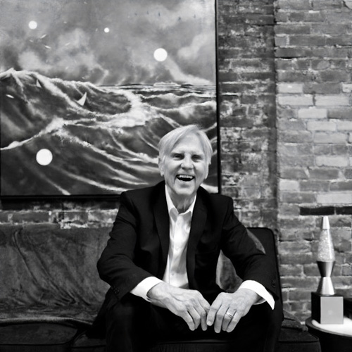 Black and white portrait of a man wearing a dark suit, sitting in front of a painting of waves and laughing