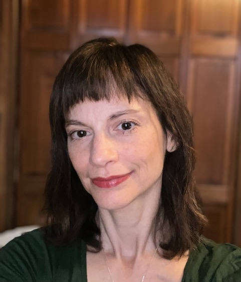 A white woman with reddish brown hair and bangs, standing in front of a wood paneled wall