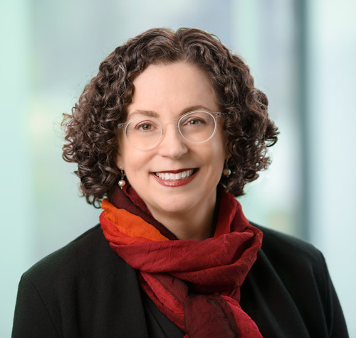 Portrait of a woman with curly brown hair, clear eyeglasses, wearing a black blazer and a red scarf