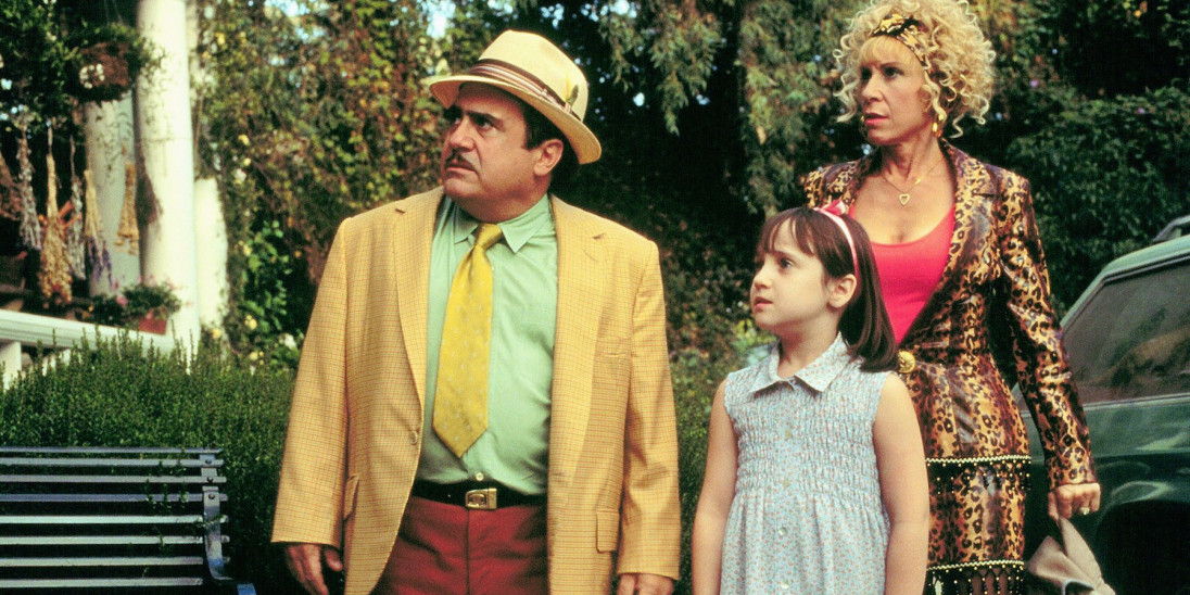 Film still of a man in a yellow suit and hat with a green shirt, a young girl in a blue sleeveless dress, and a woman in a cheetah print skirt suit looking off to the right