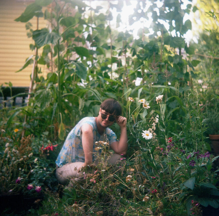 Portrait of artist Sam Tam Ham squatting down in a garden of tall plants