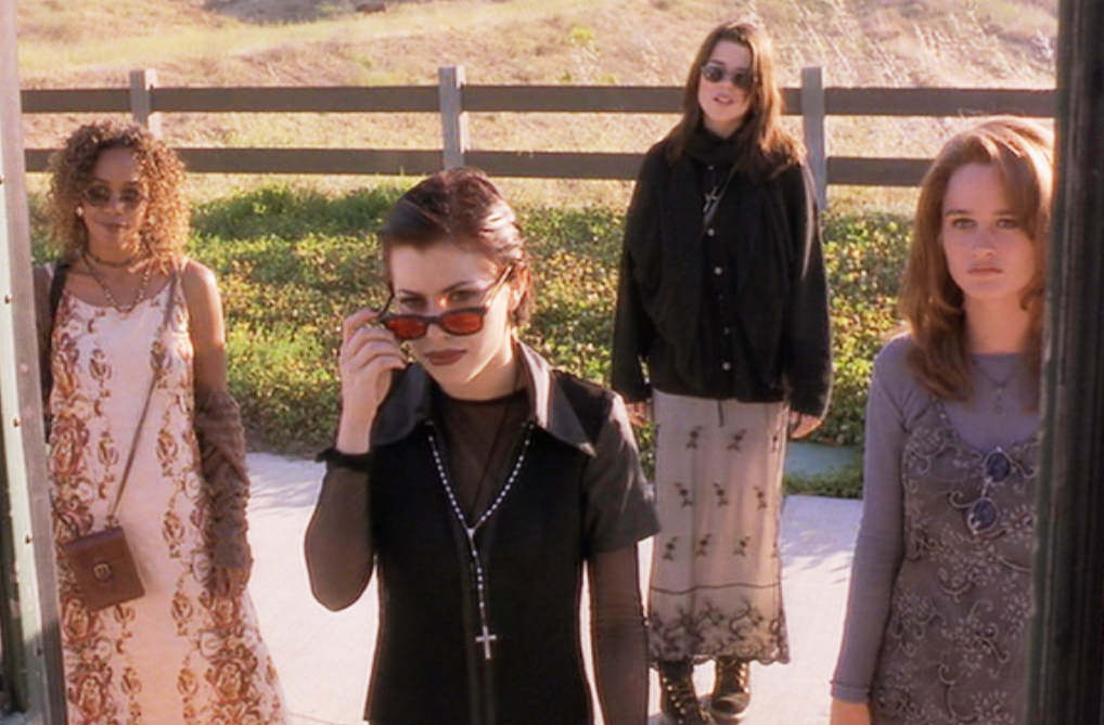 Film still of a group of four young women standing outside in front of a fence. Three are wearing sunglasses and the one in front is looking over the top of her sunglasses.