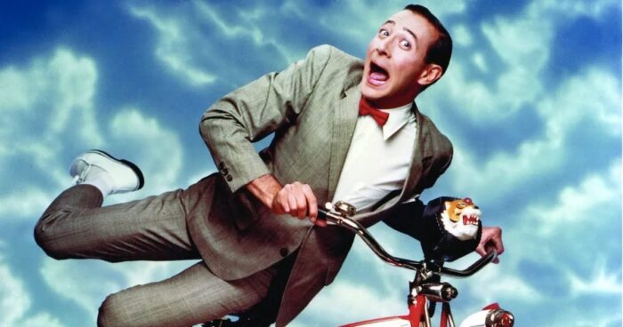 Film still of a white man with short brown hair in a grey suit, red bowtie, and white shoes holding on to a red bicycle against a blue sky with clouds