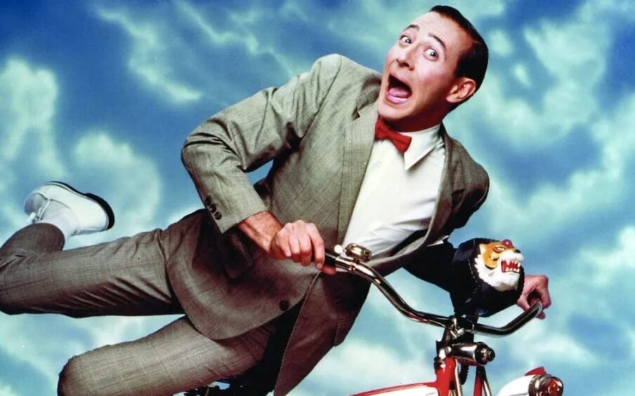 Film still of a white man with short brown hair in a grey suit, red bowtie, and white shoes holding on to a red bicycle against a blue sky with clouds