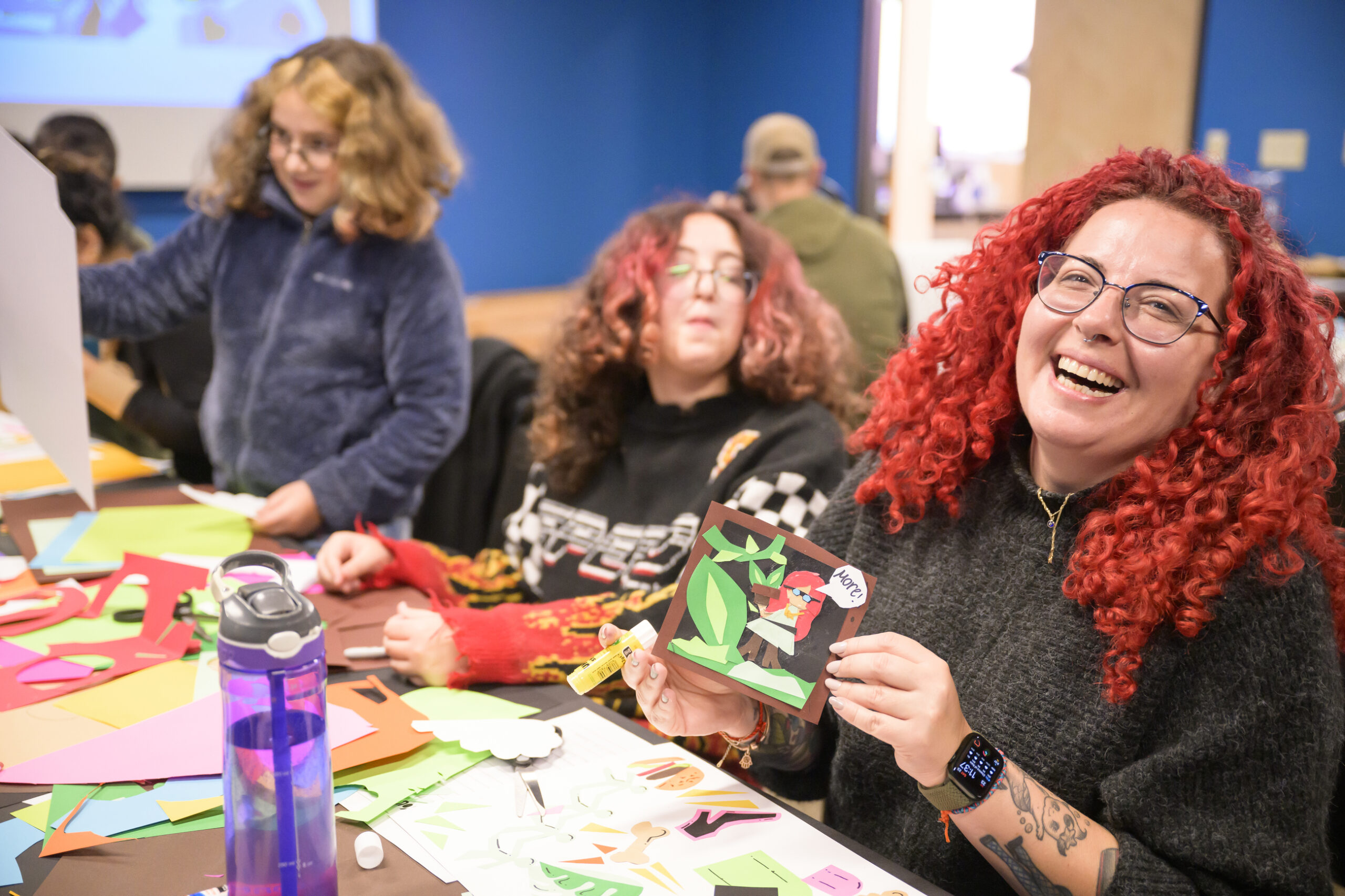 A woman with curly bright red hair and eyeglasses laughing next to her kids at a craft table