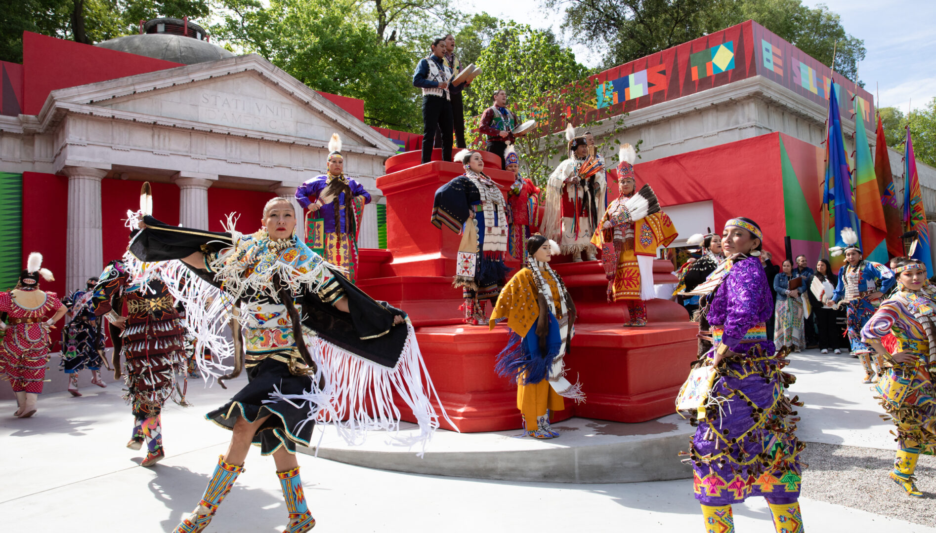 Photo of jingle dancers, people in bright, patterned dresses and boots covered in bells, dancing in front of the U.S. Pavilion in Venice, Italy.