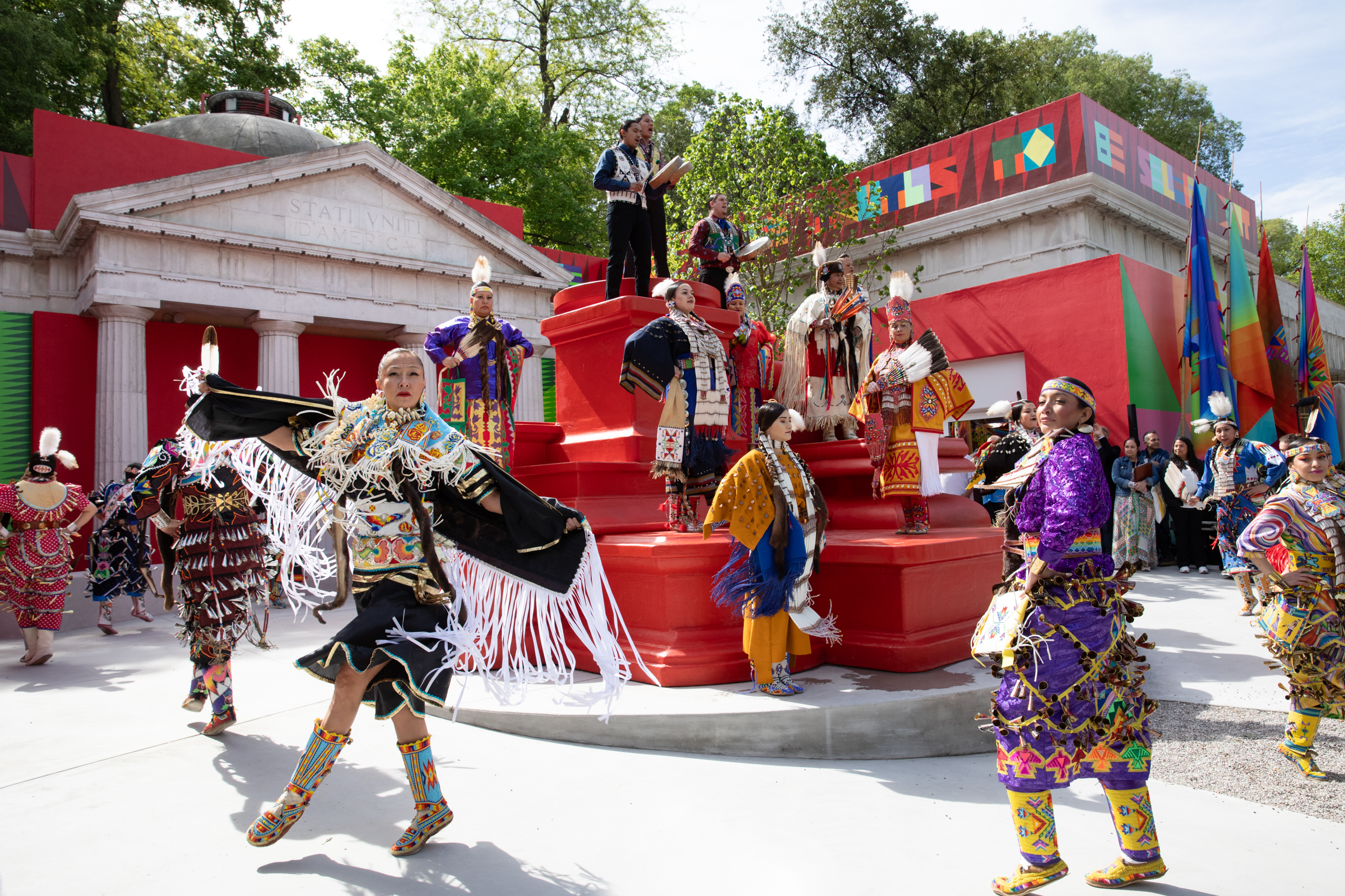 Photo of jingle dancers, people in bright, patterned dresses and boots covered in bells, dancing in front of the U.S. Pavilion in Venice, Italy.