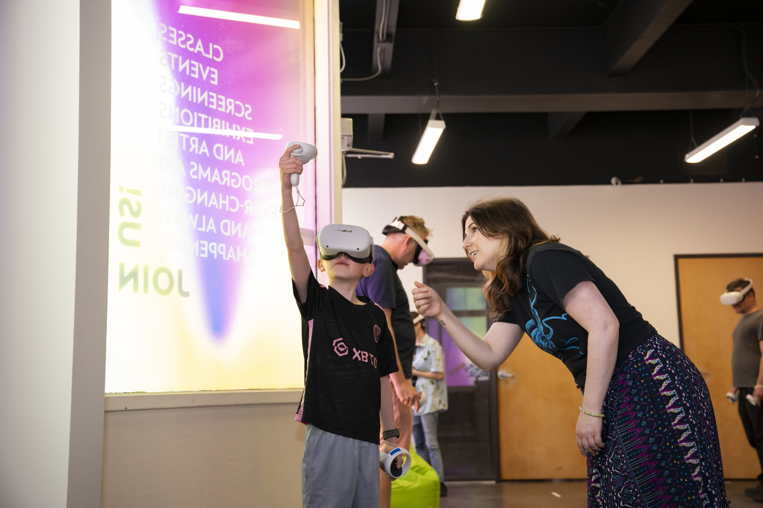 A kid in virtual reality googles and holding virtual reality controls being helped by a teacher