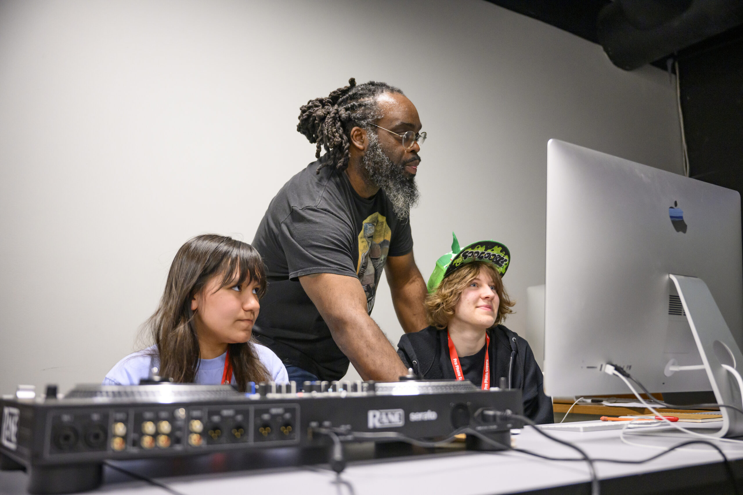 A Black man with dreadlocks working at a computer and DJ equipment witha. teenage boy and girl