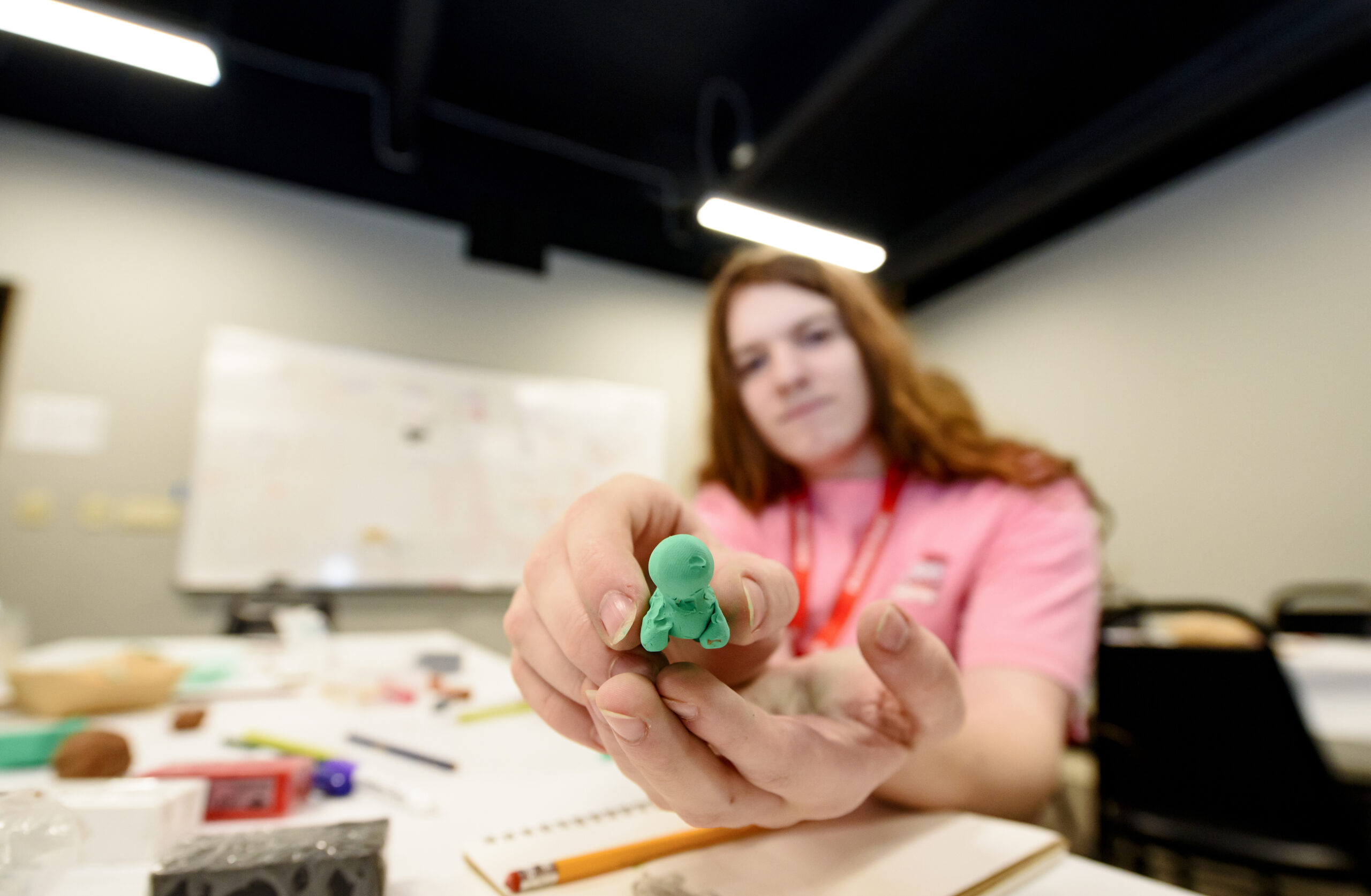A teenager with long red hair holding out a green small sculpture toward the camera