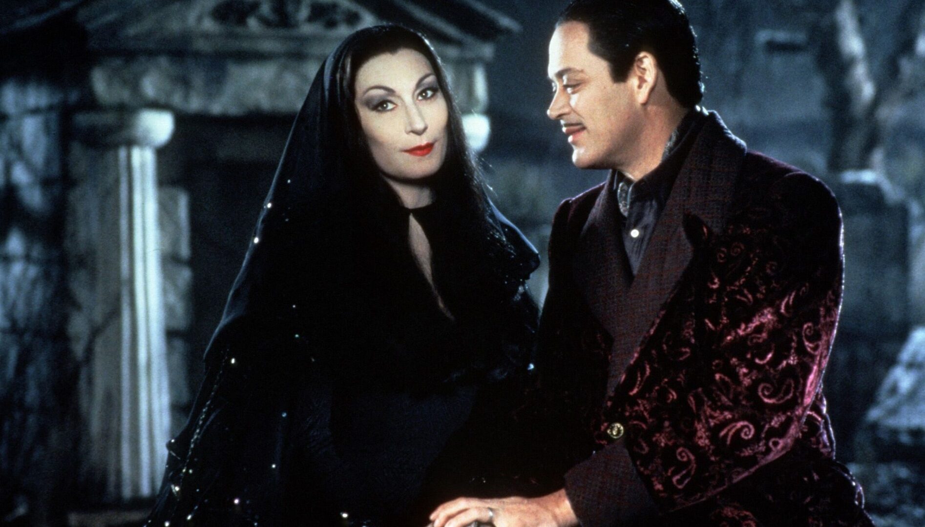 A pale woman with long black hair and dramatic eye makeup holding hands with a man with slicked-back black hair and a mustache, wearing a velvet coat