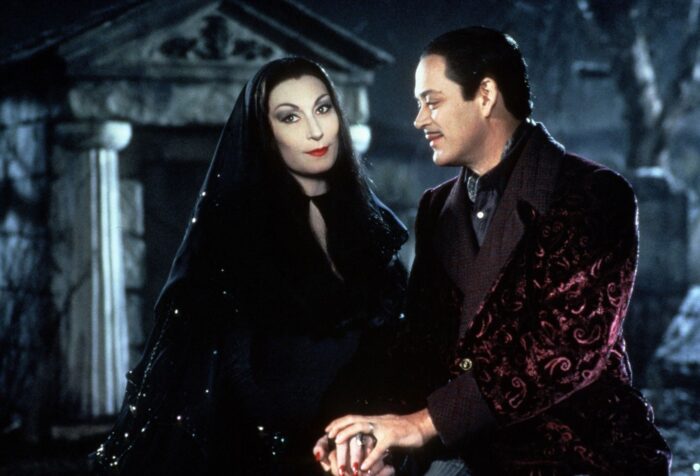 A pale woman with long black hair and dramatic eye makeup holding hands with a man with slicked-back black hair and a mustache, wearing a velvet coat