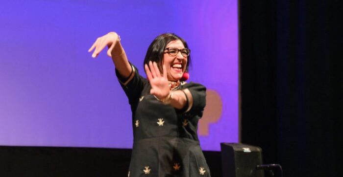 Smiling Indian woman with a brown bob haircut and eyeglasses. She's dancing on stage in front of a purple screen.