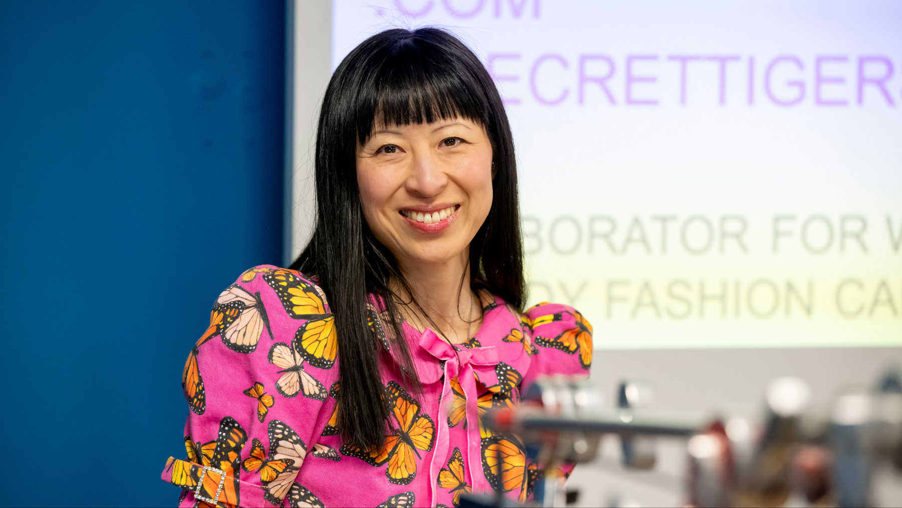 A smiling woman with long black hair and blunt bangs, wearing a bright pink dress with butterflies on it