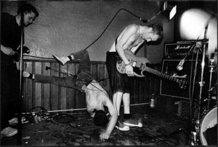 Black and white photo of a hardcore band on stage. Two are bent over guitars and one is flailing on the stage.