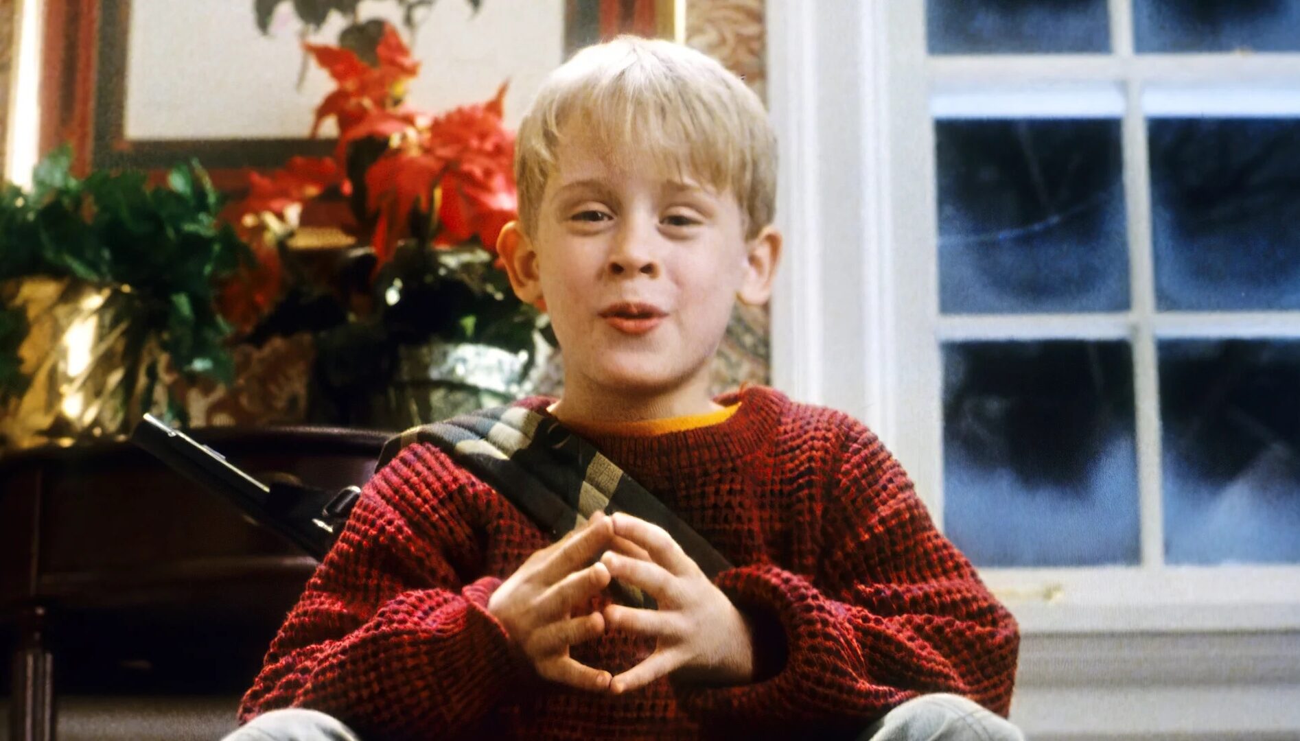 A young blond boy with a red sweater sitting with a frosty window and Christmas decorations behind him