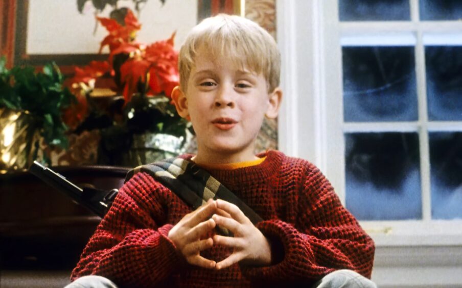 A young blond boy with a red sweater sitting with a frosty window and Christmas decorations behind him