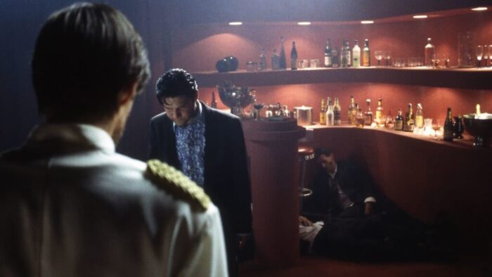 Two men standing in a dark bar. Two men are slumped against the bar.