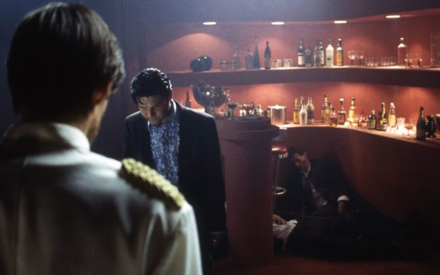 Two men standing in a dark bar. Two men are slumped against the bar.