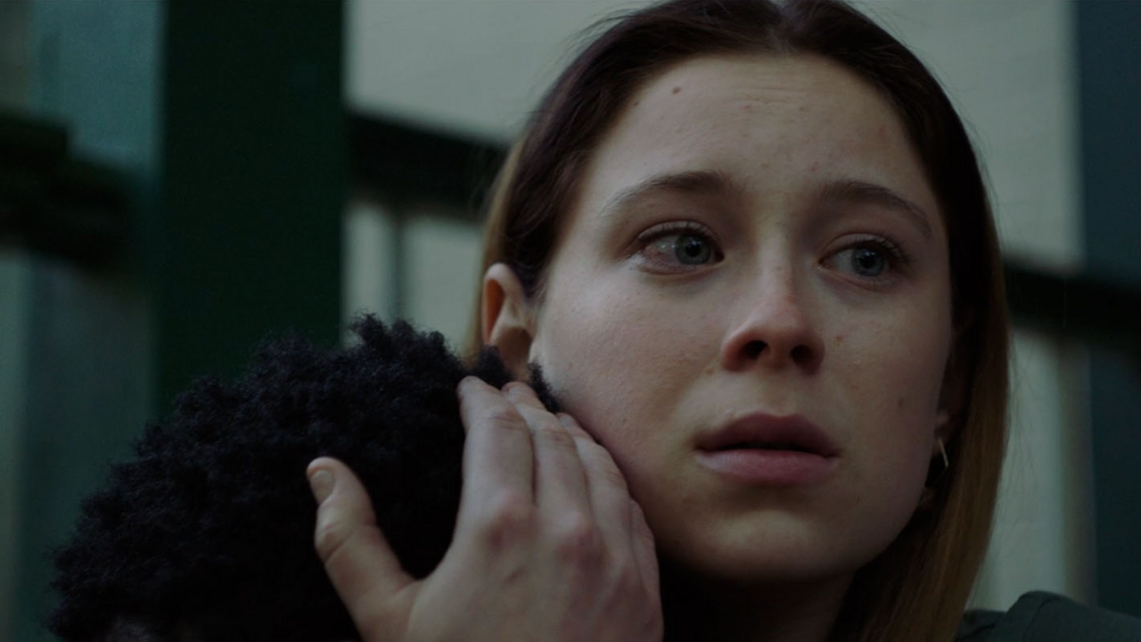 A woman with a worried look on her face hugging someone with curly dark hair whose back is to the camera