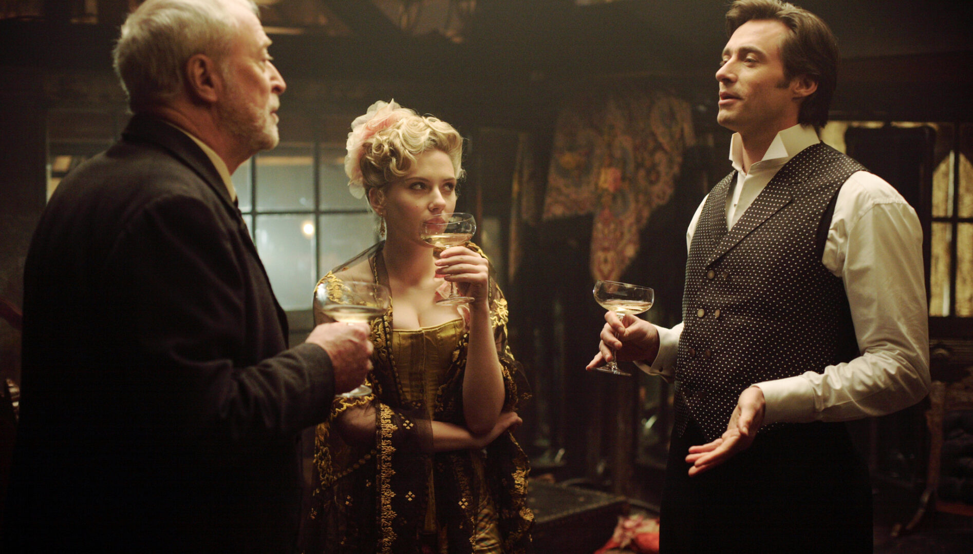 An older white man with grey hair talking and drinking with a white woman with blonde hair and a man with brown hair. All are wearing early 20th century clothing.