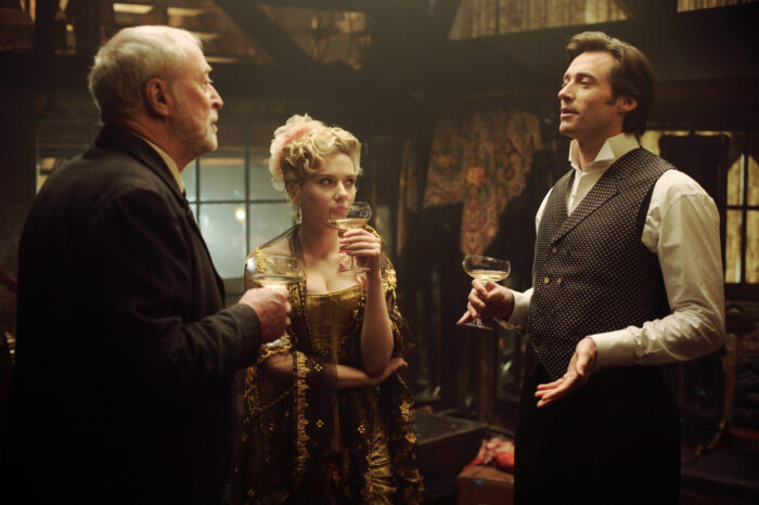 An older white man with grey hair talking and drinking with a white woman with blonde hair and a man with brown hair. All are wearing early 20th century clothing.