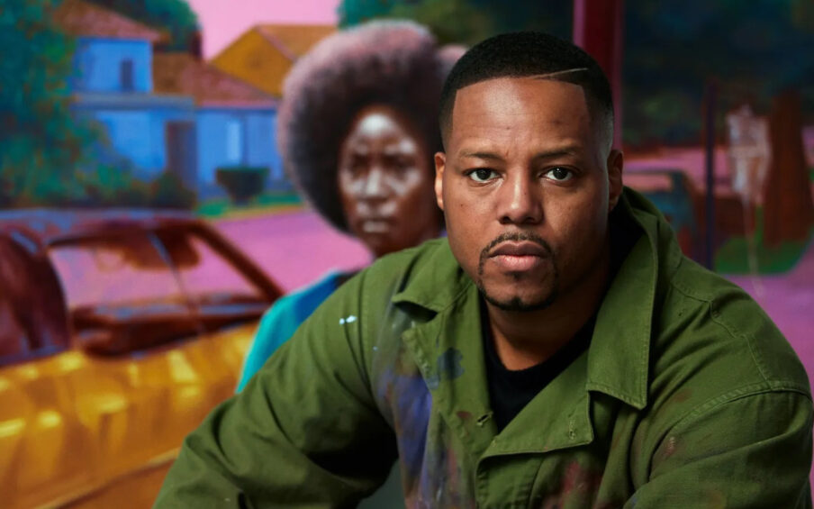 Portrait of a Black man wearing an army green jacket, sitting in front of a painting of a woman with an afro