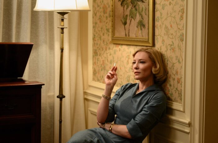 Film still of a woman in a 1950s style blue dress with blonde hair. She's leaning against patterned wallpaper near an armchair and a lamp and is smoking a cigarette.