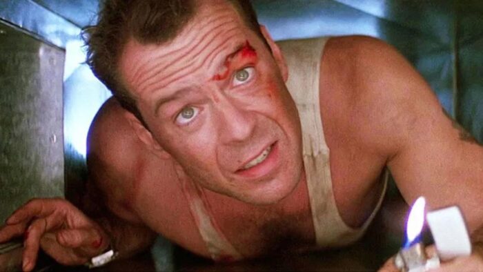 Close up of a man crawling in a vent, wearing a white sleeveless shirt, holding a lighter. He has a cut next to his eye.