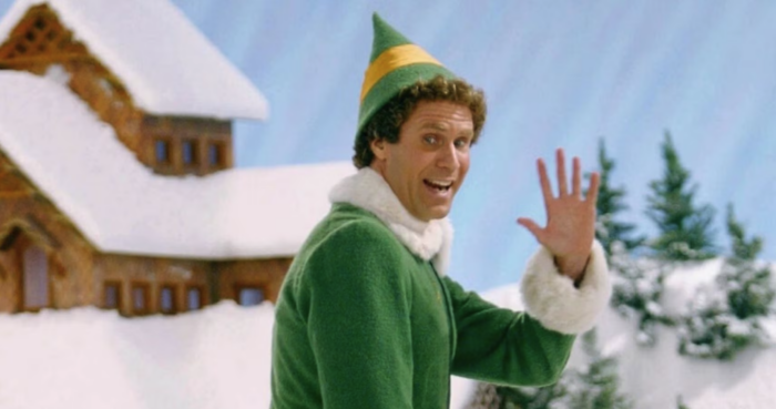 Film still of a man with curly blond hair waving, wearing a green elf costume, in a snowy outdoor scene.