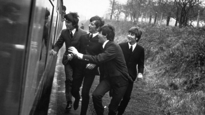 Black and white film still of The Beatles wearing suits and running alongside a train