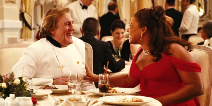 A white man with blonde hair and a white chef's jacket sitting at a restaurant table with Black woman wearing a red sleeveless gown. They are smiling and turned towards each other.