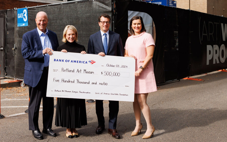 Photo of four smiling people, holding a big check in front of a construction site