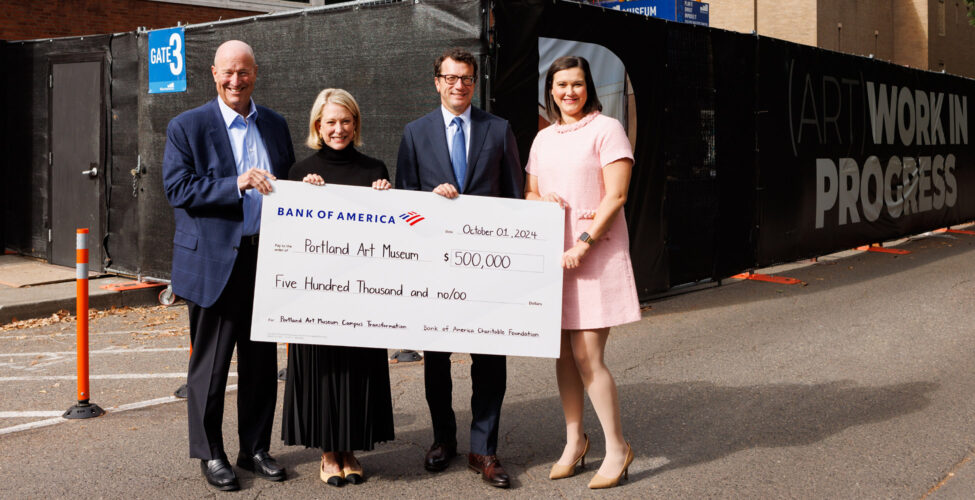 Photo of four smiling people, holding a big check in front of a construction site