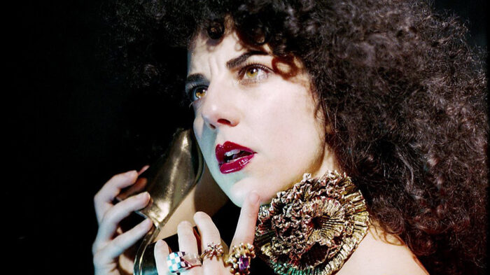 Portrait of a woman with long brown curly hair, wearing red lipstick, a blue sleeveless top, a watch, multiple rings, and holding up a gold high heel to her ear