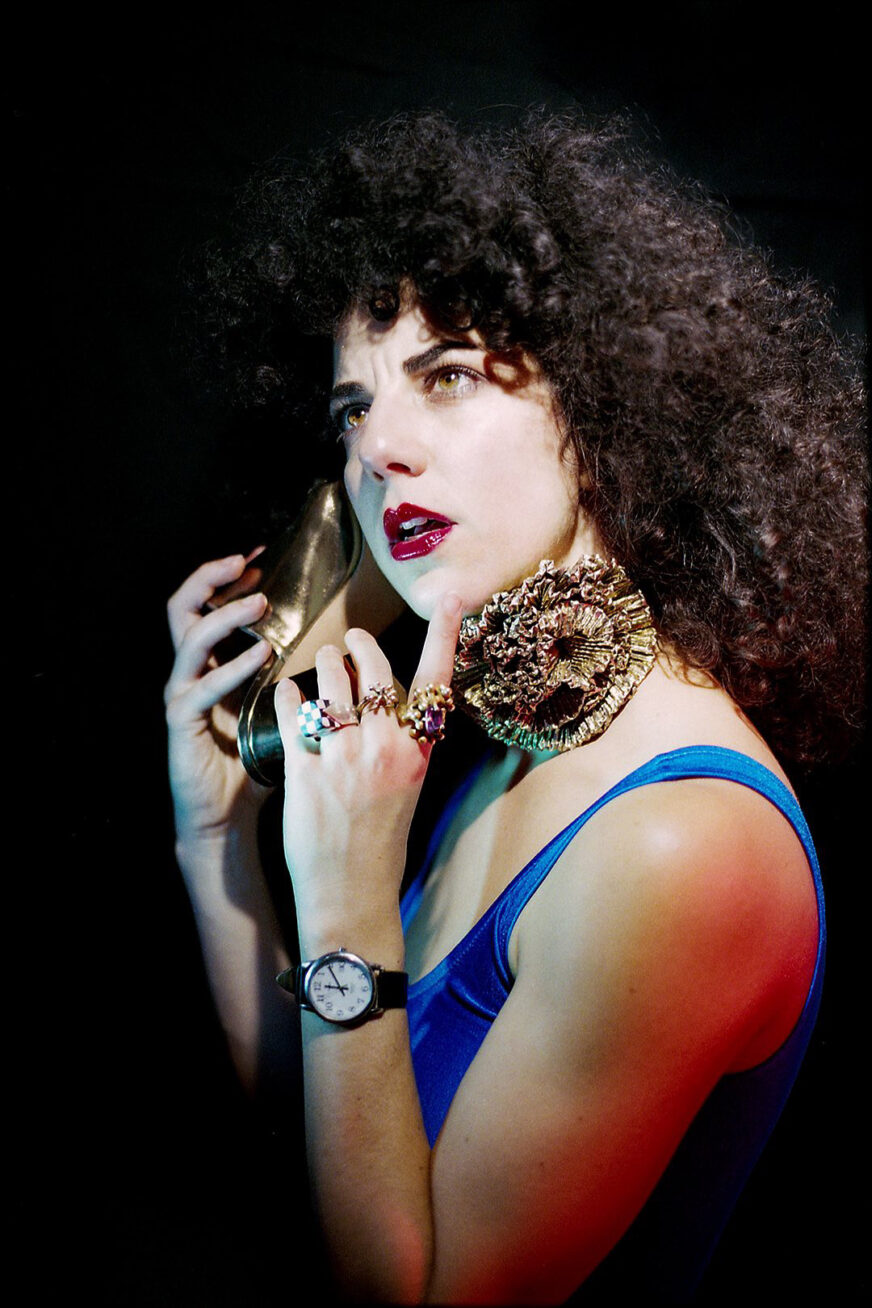 Portrait of a woman with long brown curly hair, wearing red lipstick, a blue sleeveless top, a watch, multiple rings, and holding up a gold high heel to her ear