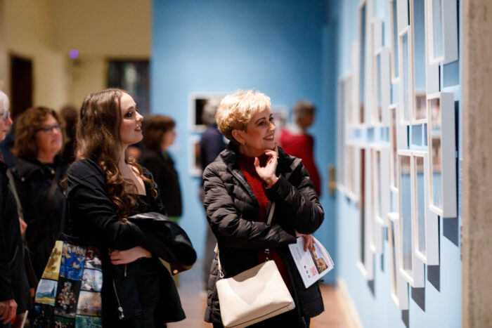 A smiling woman looking at photographs hanging in white frames on a light blue wall