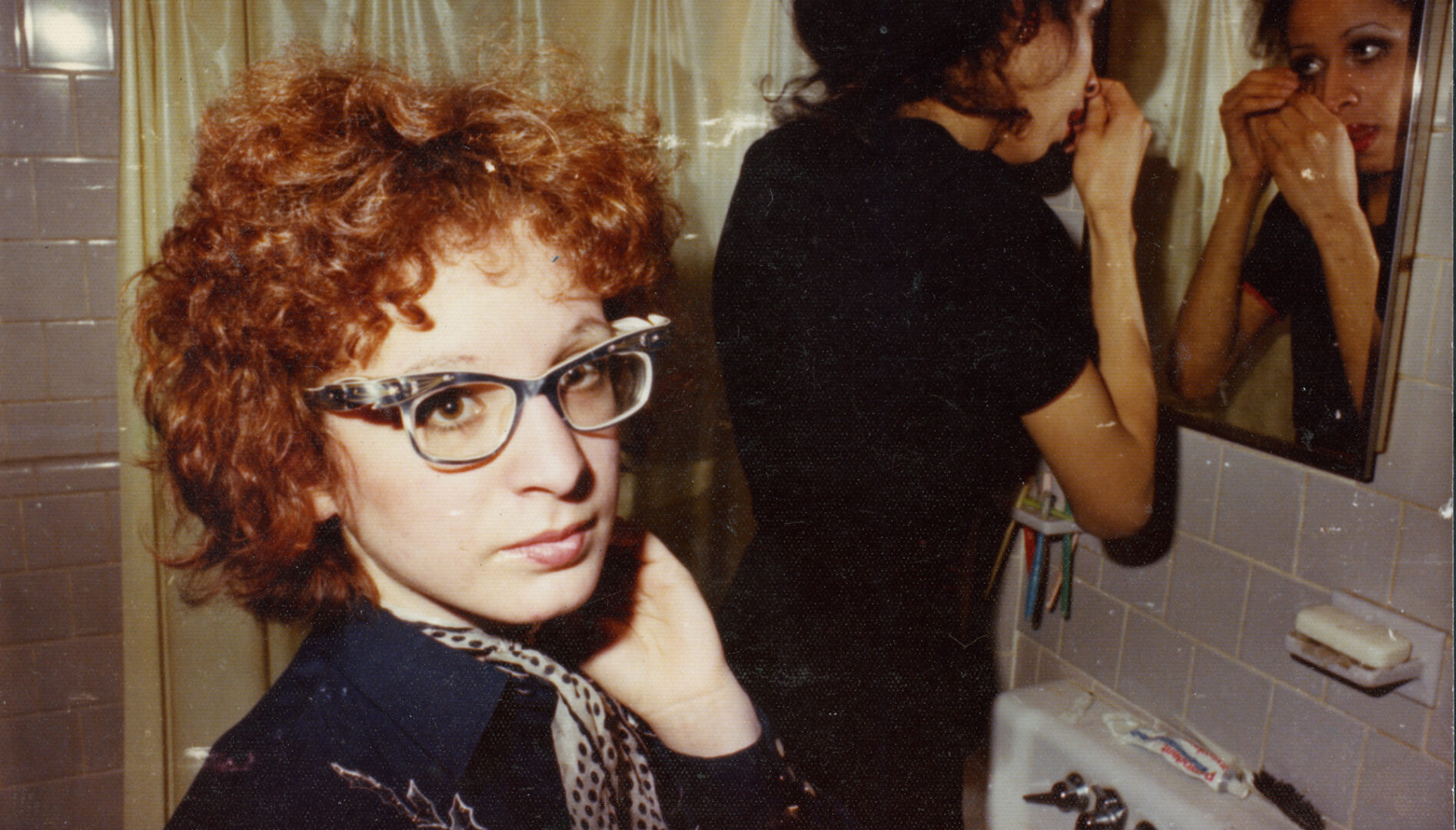 A woman with red, curly hair and cat's eye glasses stands in a bathroom with another woman looking in a mirror