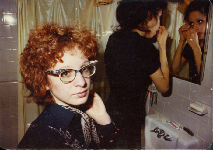 A woman with red, curly hair and cat's eye glasses stands in a bathroom with another woman looking in a mirror