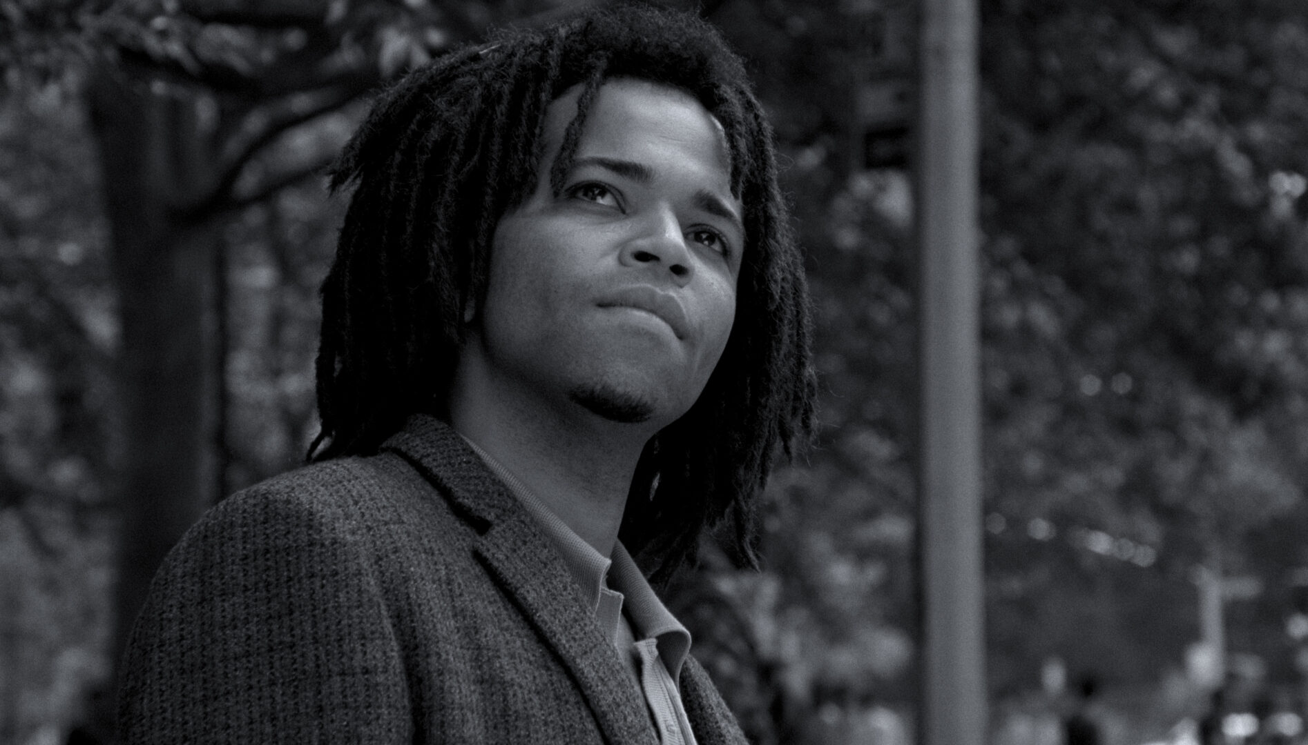 A black and white film still of a young Black man with dreadlocks, wearing a suit jacket, looks up towards the sky