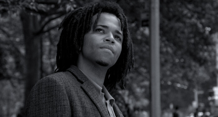 A black and white film still of a young Black man with dreadlocks, wearing a suit jacket, looks up towards the sky