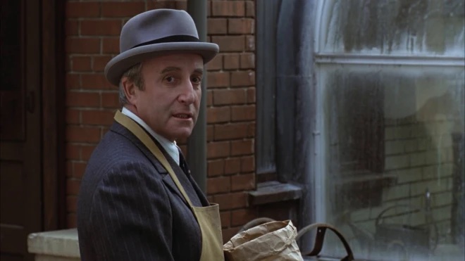 An older man in a grey bowler hat, a suit, and an apron is turning towards the camera in front of a brick wall and window